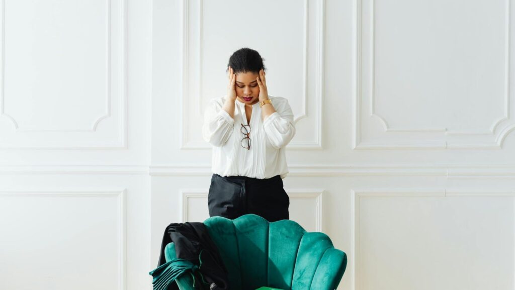A blank woman wearing a white blouse stands behind a small green velvet chair. She is clutching her face with both hands and looking down in distress.