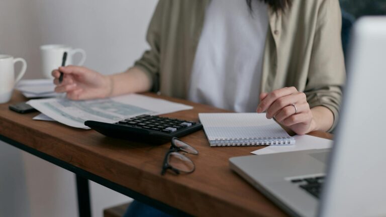 A woman sits at a table will bills, a computer, and a calculator. She has a pen in her hands and you cannot see her face.