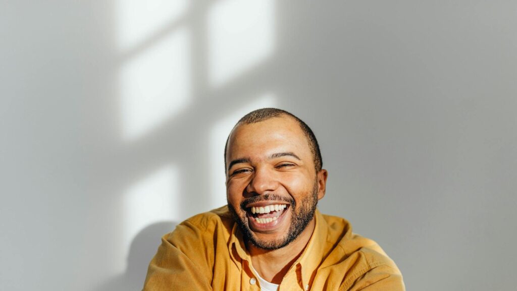 A black man in a yellow hoodie stands in front of a white wall smiling with the sun hitting his face.