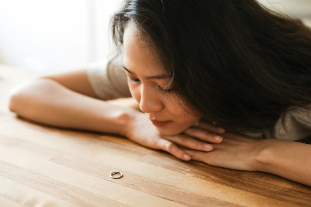 A woman with long, dark hair rests her chin on the back of her hands as she slumps down on a table, looking at a wedding ring she is not wearing.