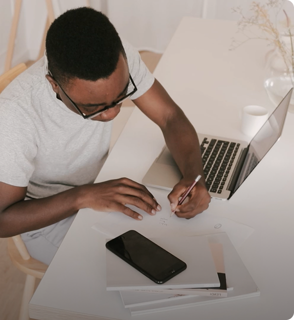 Person writing notes at a desk with a laptop and phone